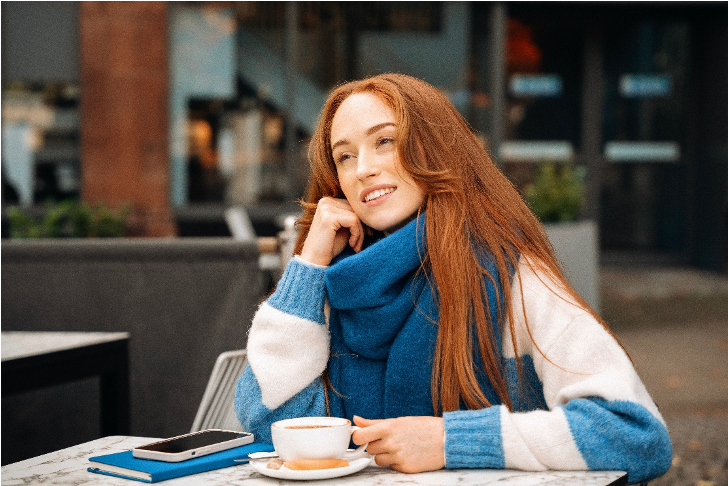 Young woman enjoying coffee at an outdoor city café on a sunny winter day, wearing a warm sweater and scarf